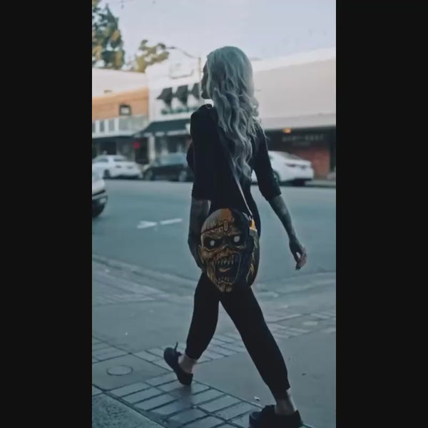 video, A blond woman dressed in black, walking while carrying Iron Maiden purse. eddie face, yellow blue red and black ,blood coming from middle of forehead large black eyes, open scowling mouth showing teeth and tongue